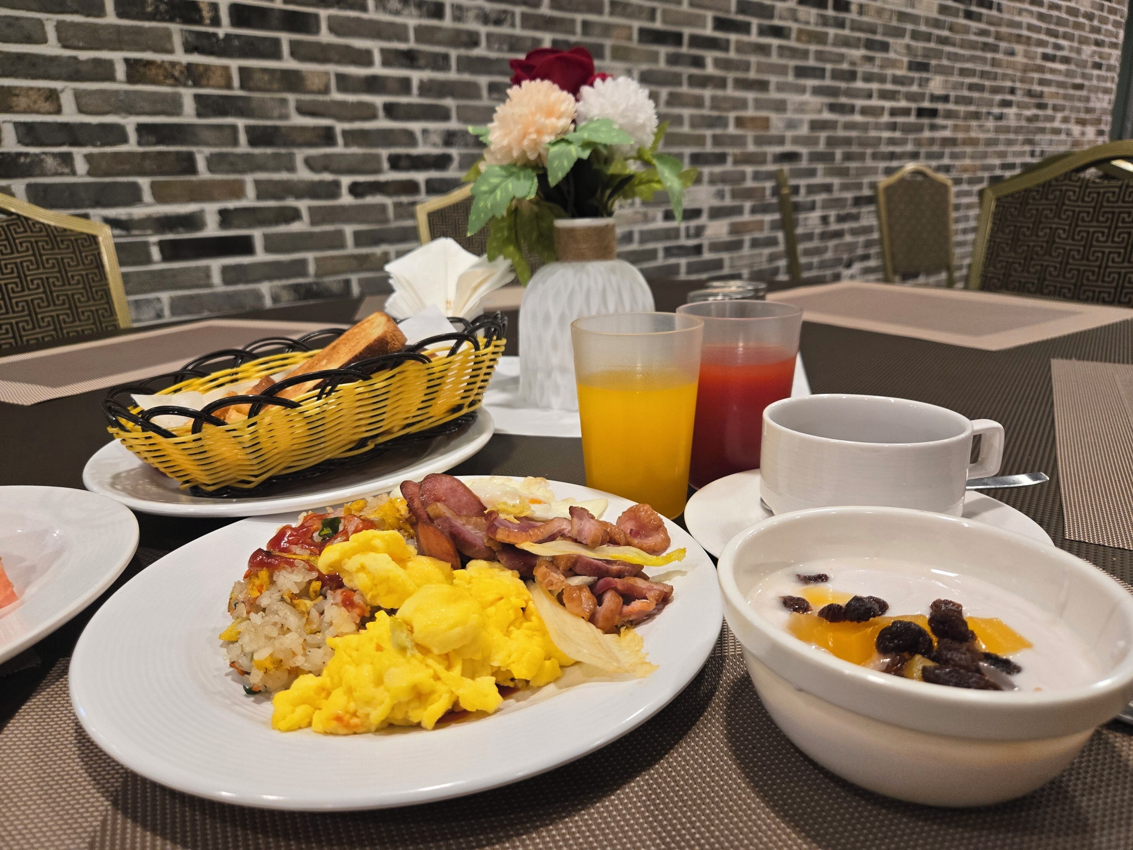 Desayuno con huevos revueltos, tocino, arroz frito, un vaso de jugo de naranja, una taza de café y un tazón de cereal con pasas.