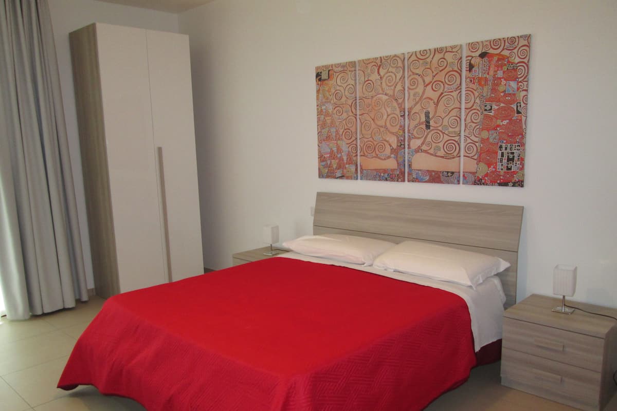A bedroom with a large bed, a nightstand, a wardrobe, and a wall with a colorful painting.