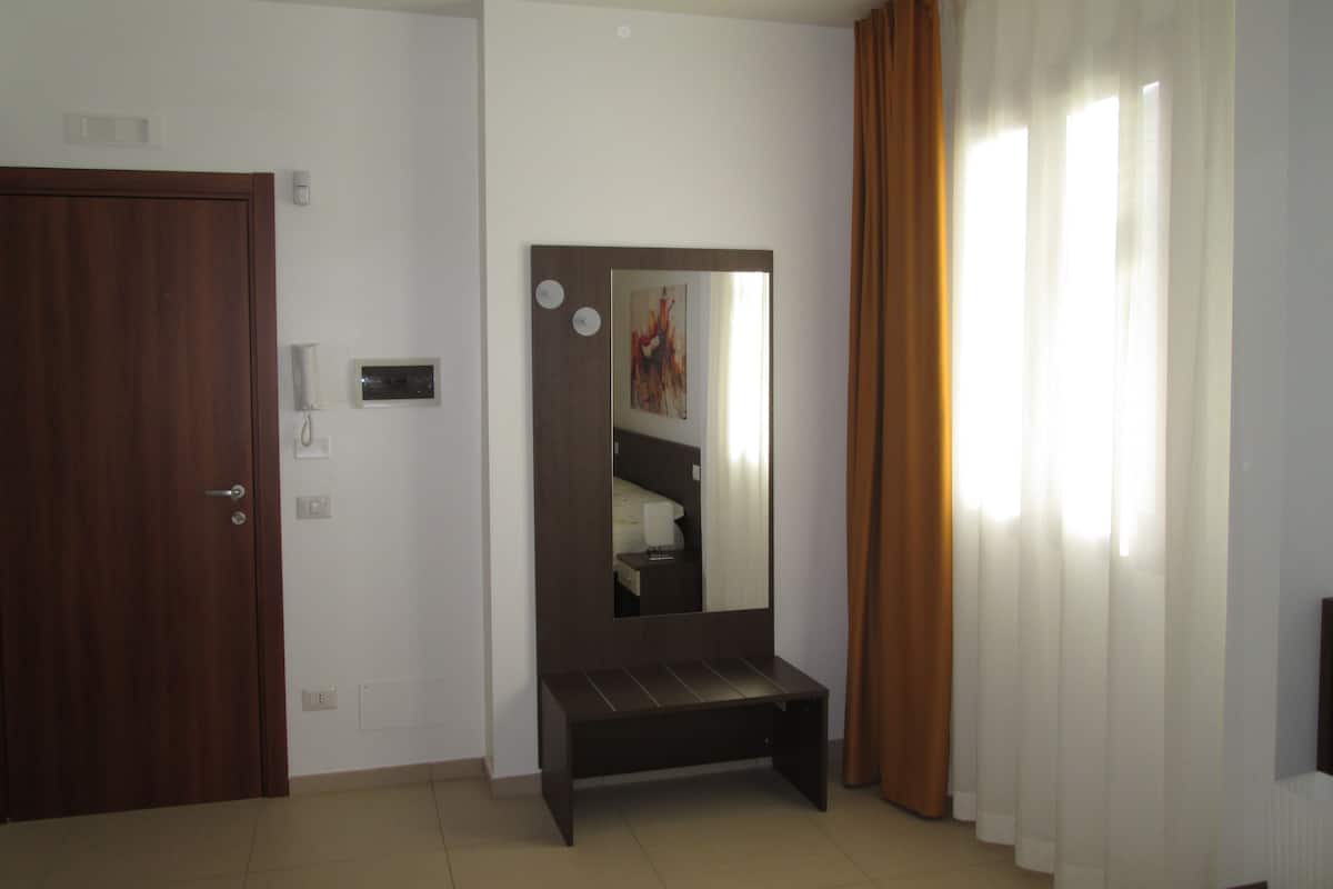 A hotel room with a wooden door, a bedside table, a mirror, a bed, and a window with curtains.