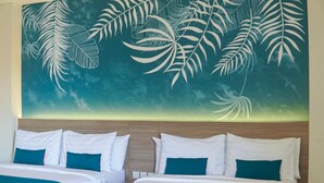 Habitación de hotel con dos camas, ropa de cama blanca, detalles en color aguamarina y un mural grande con hojas tropicales.