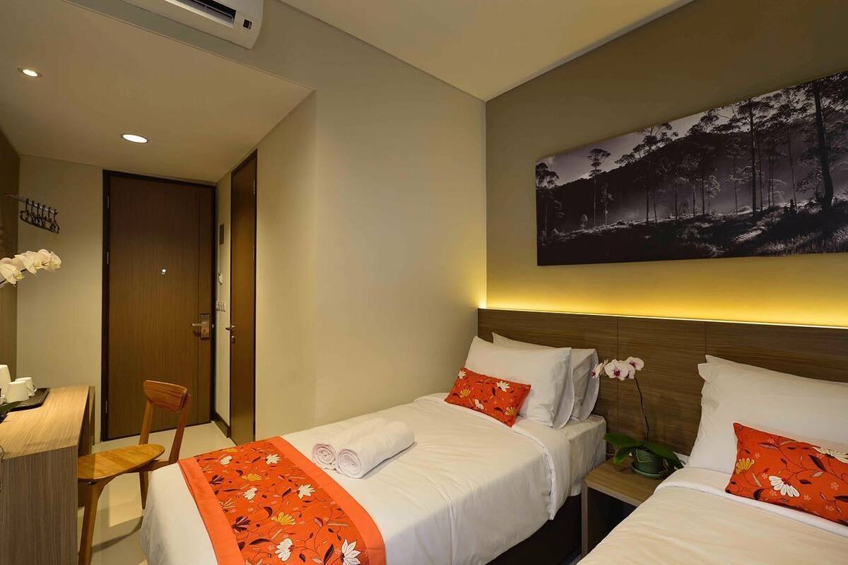 Ein Hotelzimmer mit zwei Betten, einem Schreibtisch und einem Stuhl. An der Wand hängt ein Gemälde und auf dem Nachttisch steht eine Zimmerpflanze.