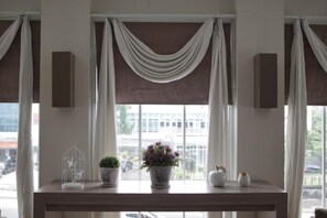 A room with a large window, draped curtains, a wooden console table with decorative items, and a view of buildings outside. - Kytos Hotel (Bandung)