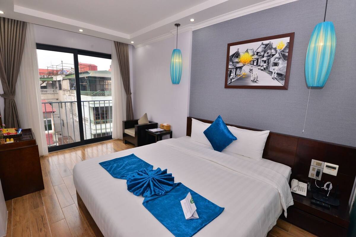 A hotel room with a large bed, a desk, a chair, a balcony with a view, and a painting on the wall.