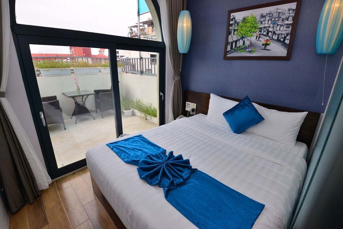 Habitación de hotel con cama, vista a través de un ventanal a un espacio exterior, un cuadro en la pared y un cojín decorativo azul.