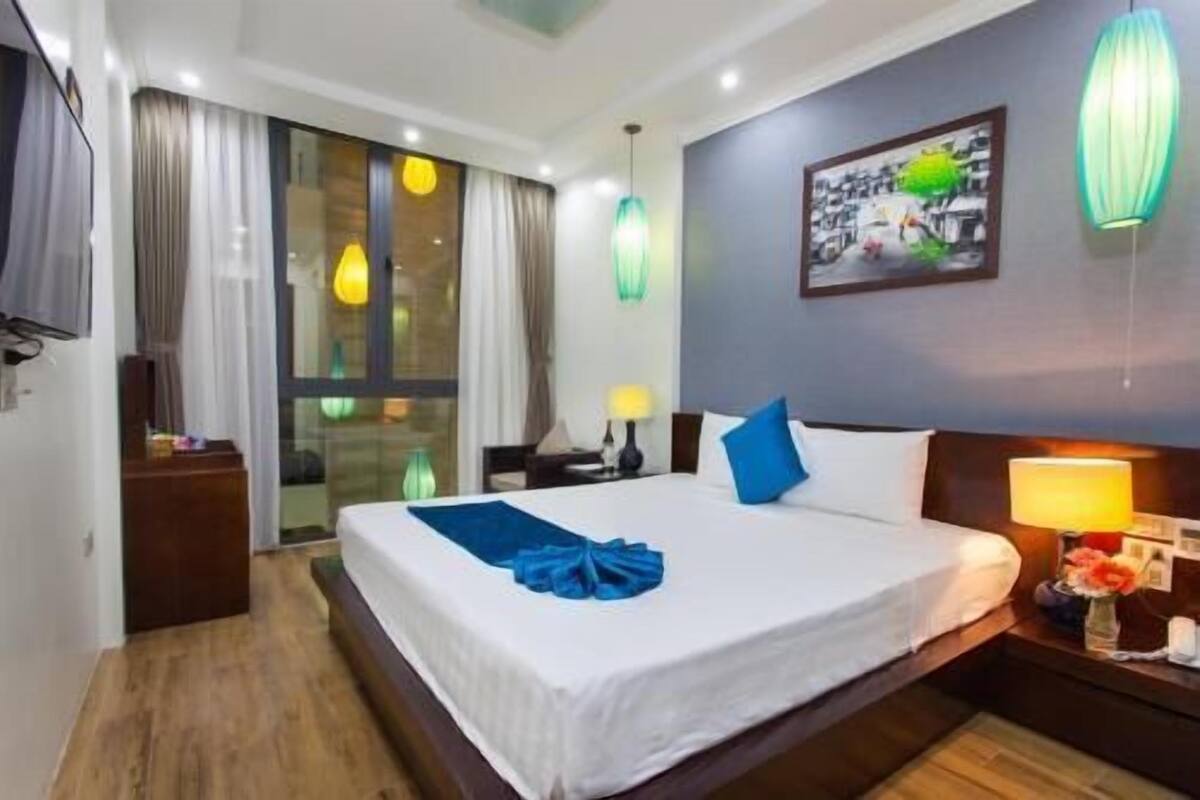 A hotel room with a bed, a nightstand, a lamp, a painting, and a window with curtains.