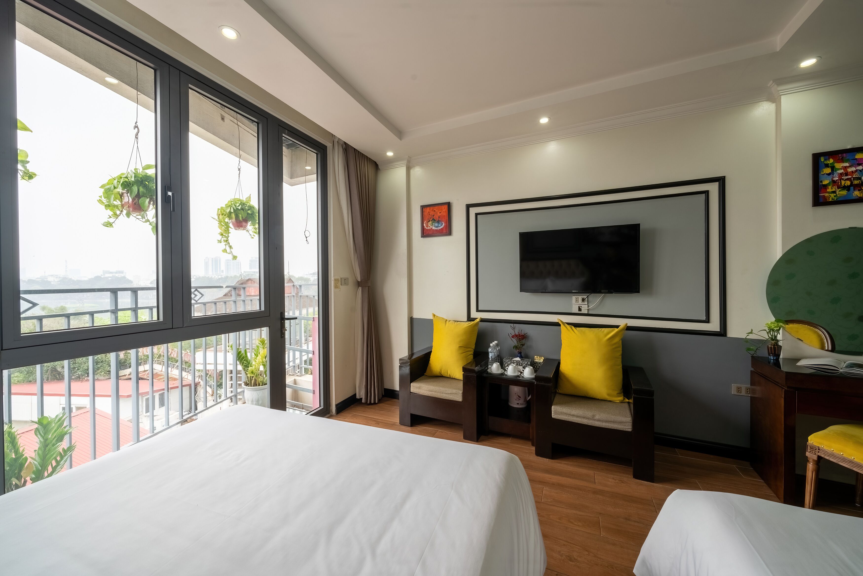 A hotel room with a large bed, two armchairs with yellow cushions, a desk, a TV mounted on the wall, and a balcony with potted plants.