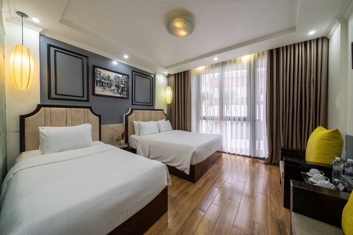 A hotel room with two beds, wooden flooring, a desk with a chair, and a window with curtains.