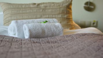 Una cama bien hecha con toallas blancas enrolladas y una decoración de hoja verde, un cojín beige, y una lámpara fijada a la pared.