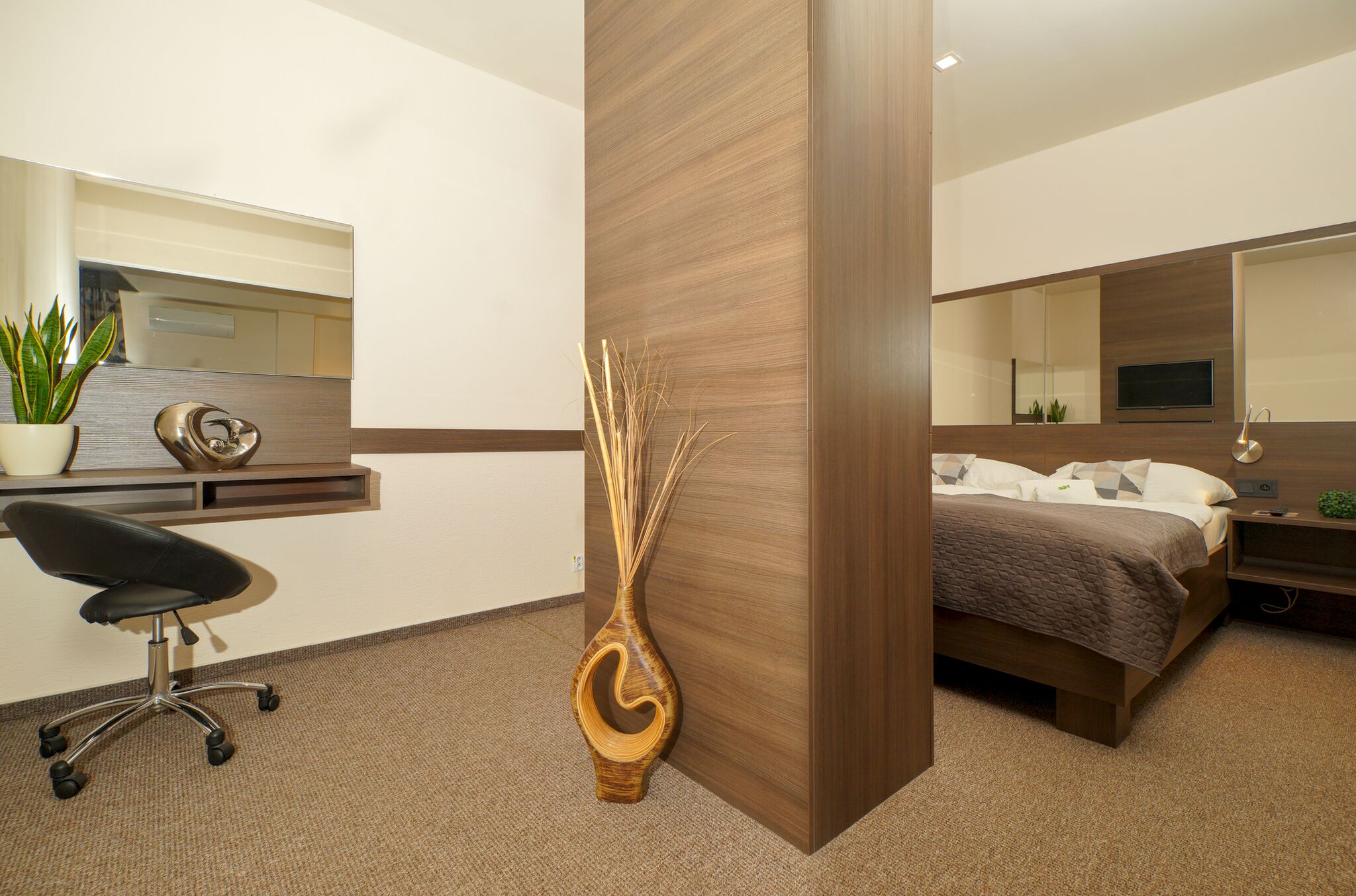 Una habitación de hotel moderna con una cama, un escritorio con una silla, un espejo grande y un jarrón decorativo.