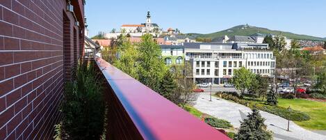 Un balcon bordé de rouge, d’où l’on peut admirer un paysage urbain avec ses bâtiments et une colline surmontée d’une tour.