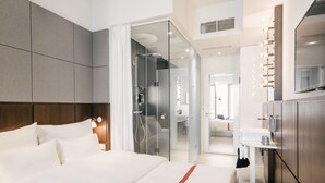 Moderni hotellihuone, jossa on suuri sänky, lasinen suihkuseinä, peilillä varustettu meikkipöytä ja ämpäri.