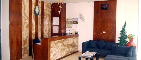 A reception area with a wooden reception desk, a blue sofa, a small table with a newspaper, a decorative fan, and a wall-mounted clock.