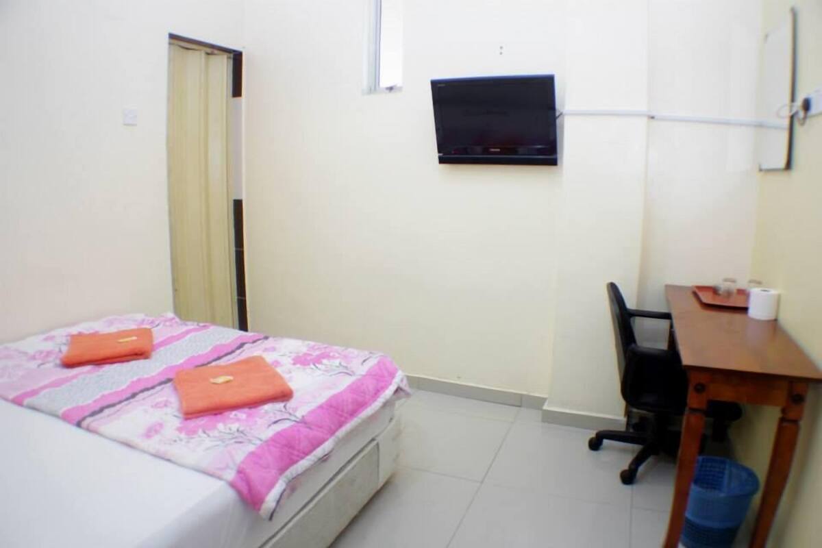 Quarto pequeno e limpo com uma cama, uma escrivaninha, uma cadeira, uma televisão e uma janela.