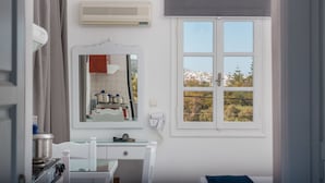 A bathroom with a white vanity, a mirror, a window with a view of buildings and trees, and an air conditioning unit. - Ktima Bianco (Naxos)