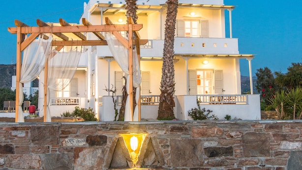 A two-story white building with a stone wall in front, a wooden pergola, and two palm trees. - Ktima Bianco (Naxos)