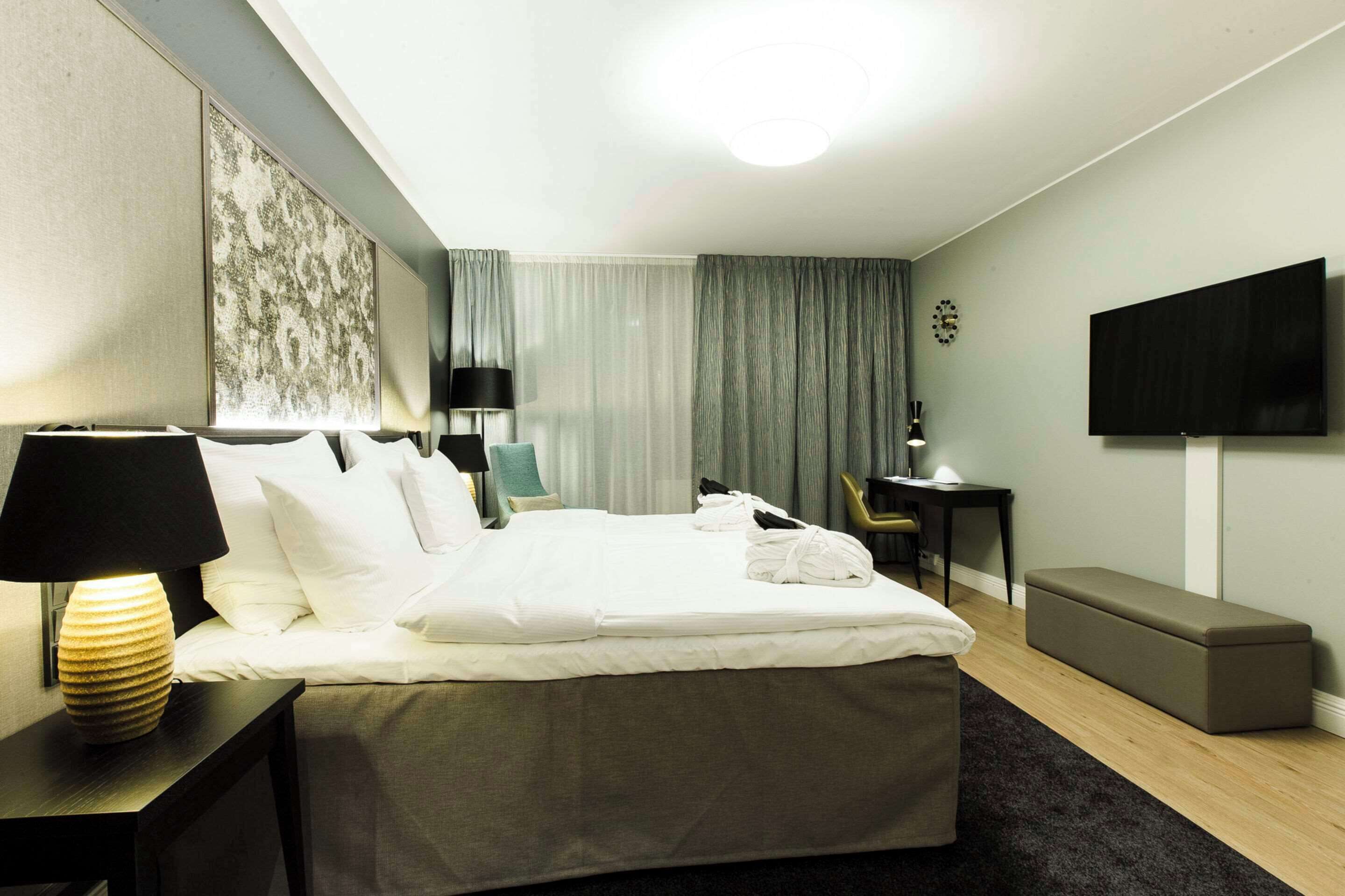 A hotel room with a bed, bedside tables, a desk, a chair, a TV, and a bench.