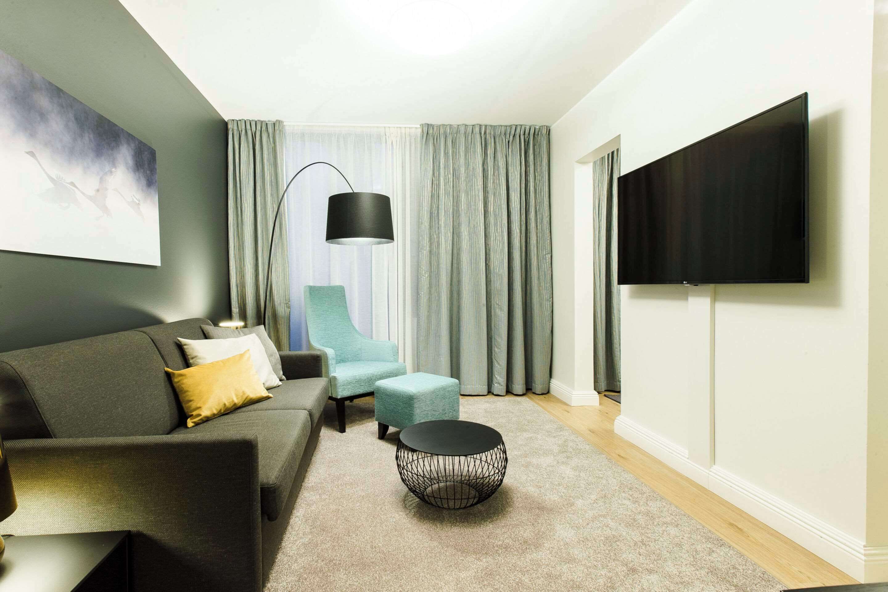 A modern living room with a grey sofa, teal armchair, and ottoman, a round black coffee table, a floor lamp, and a flat-screen TV mounted on the wall.