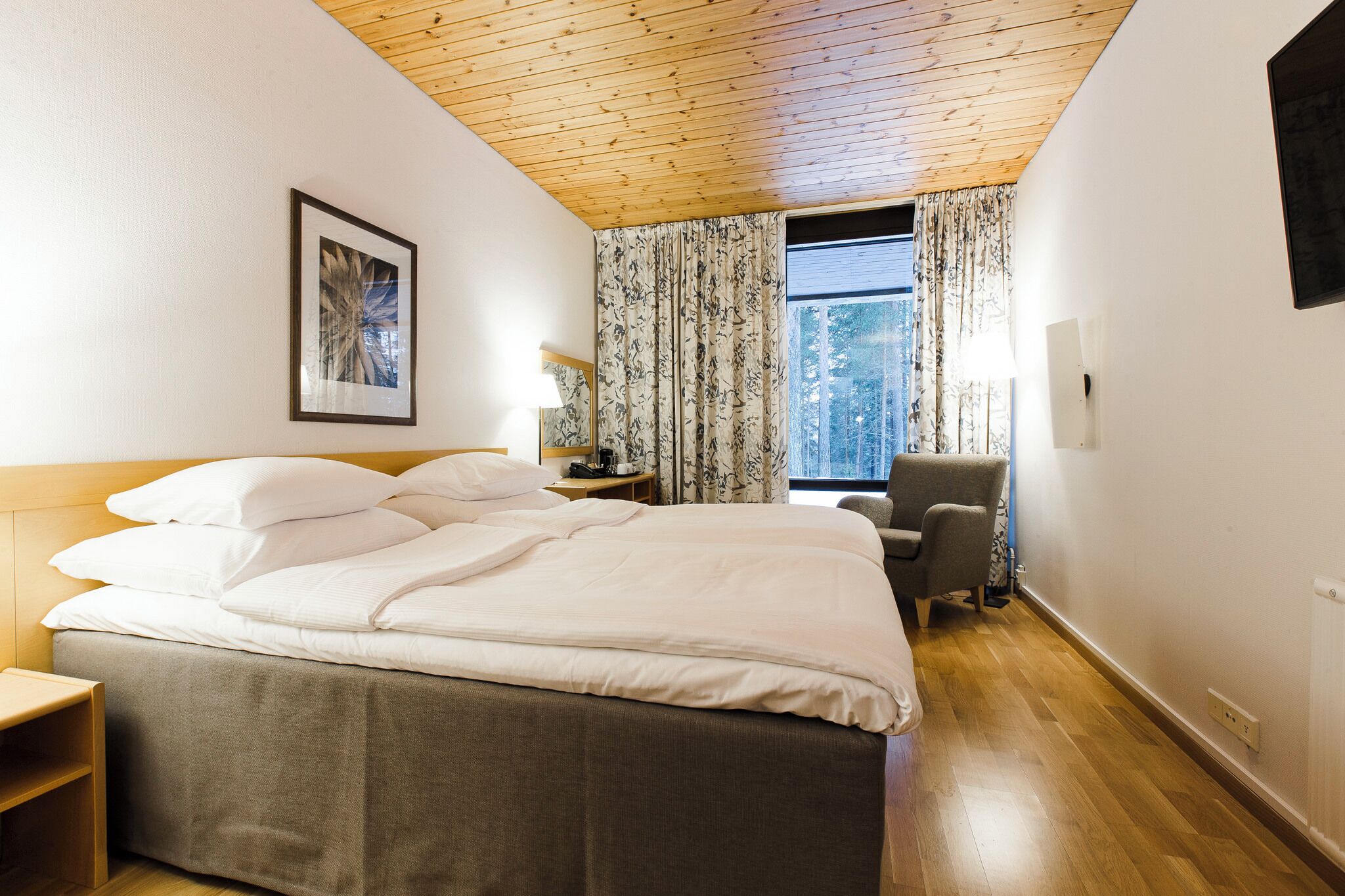 A hotel room with a wooden ceiling, a large bed with white bedding, a chair, a nightstand, and a window with curtains.