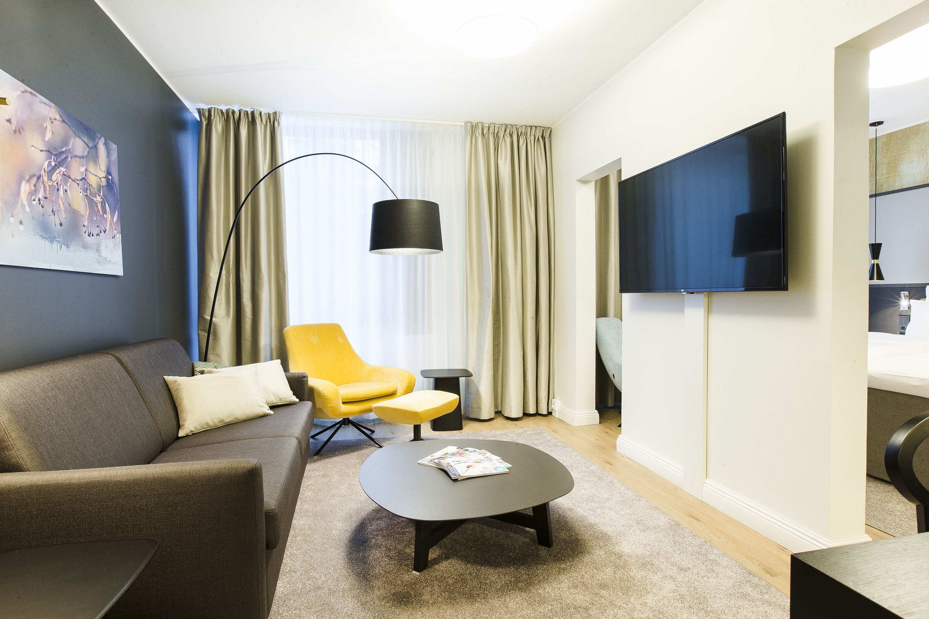A modern hotel room with a grey sofa, a yellow armchair, a small round table, and a flat-screen TV mounted on the wall.