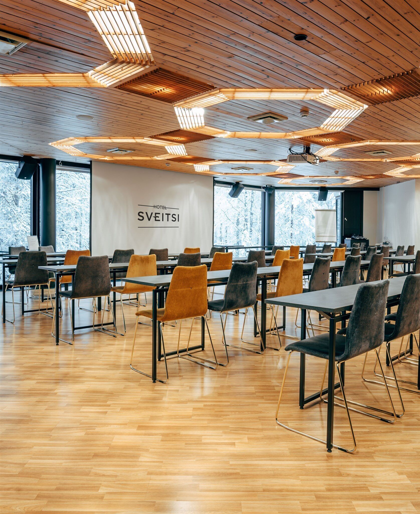A conference room with wooden floors and tables, equipped with chairs and a projector screen.
