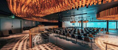 A spacious conference room with tiered seating, a wooden ceiling design, and a large screen at the front.