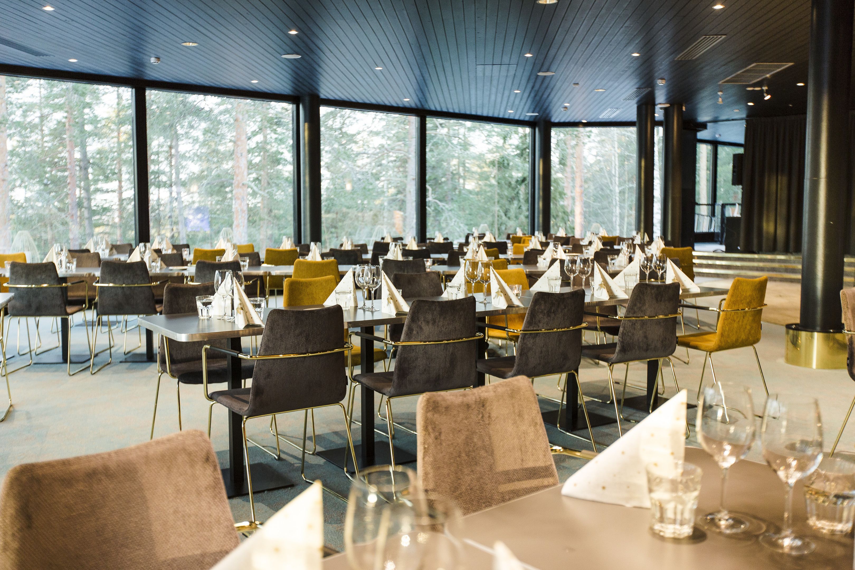 Un comedor amplio con mesas dispuestas para varios invitados, ventanas grandes que ofrecen vistas del bosque circundante y un diseño de interiores moderno.