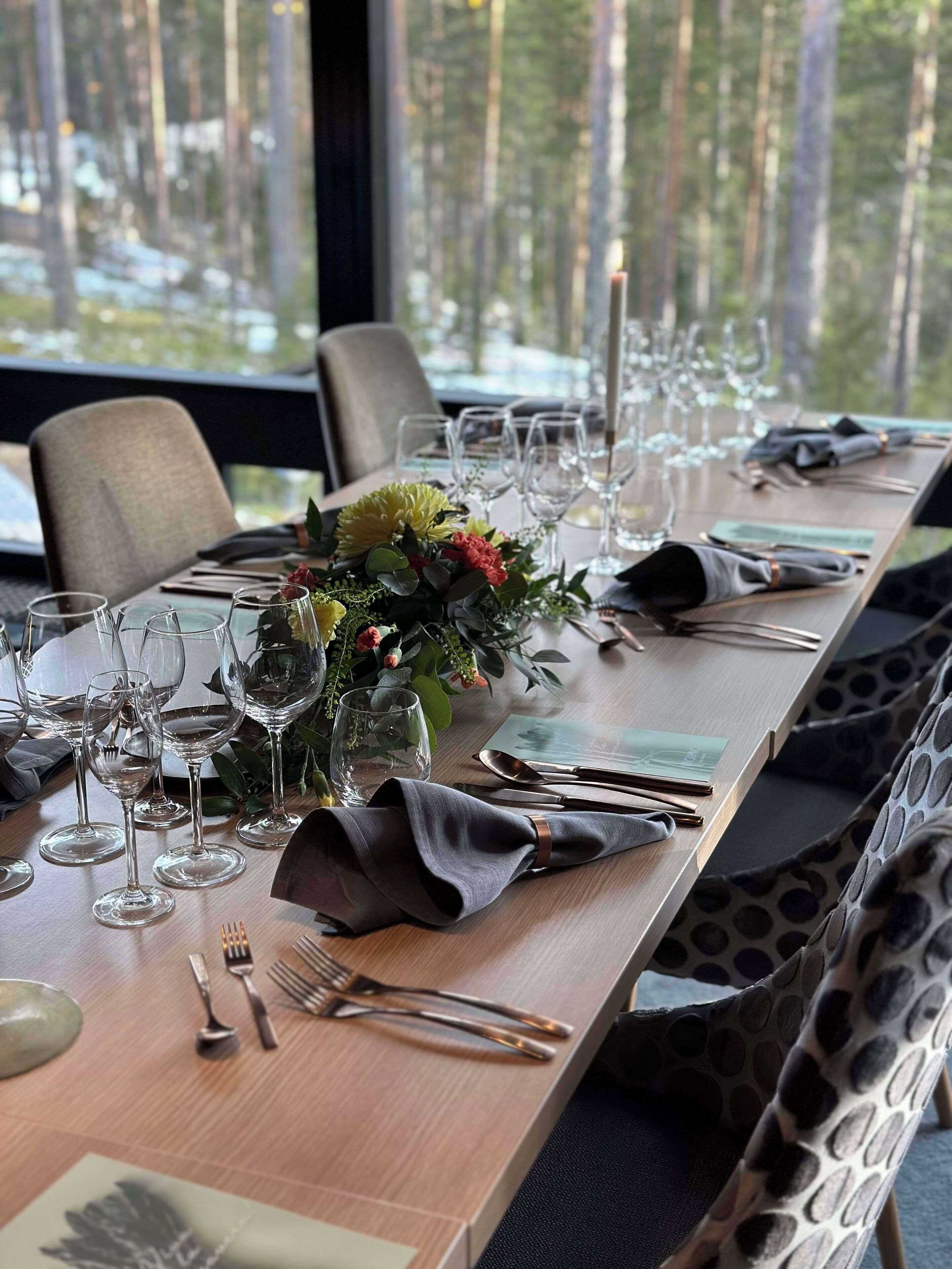 Ein Esstisch, gedeckt für eine Mahlzeit, mit Weingläsern, Besteck und einem Blumenarrangement in der Mitte.