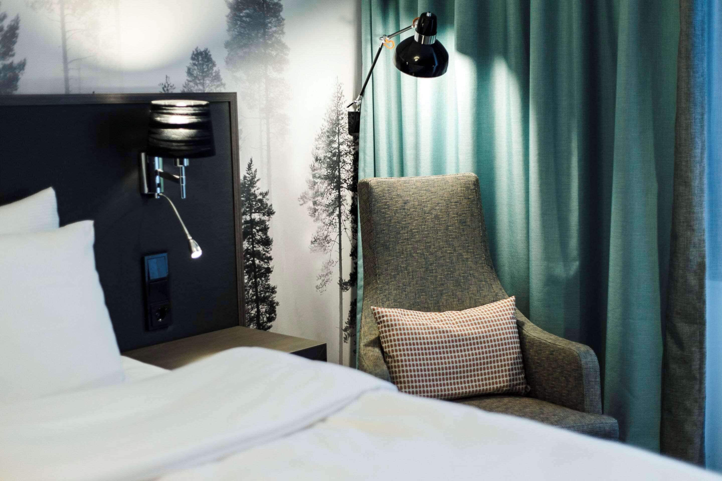 A hotel room with a bed, a chair with a pillow, a wall-mounted lamp, and a curtain.