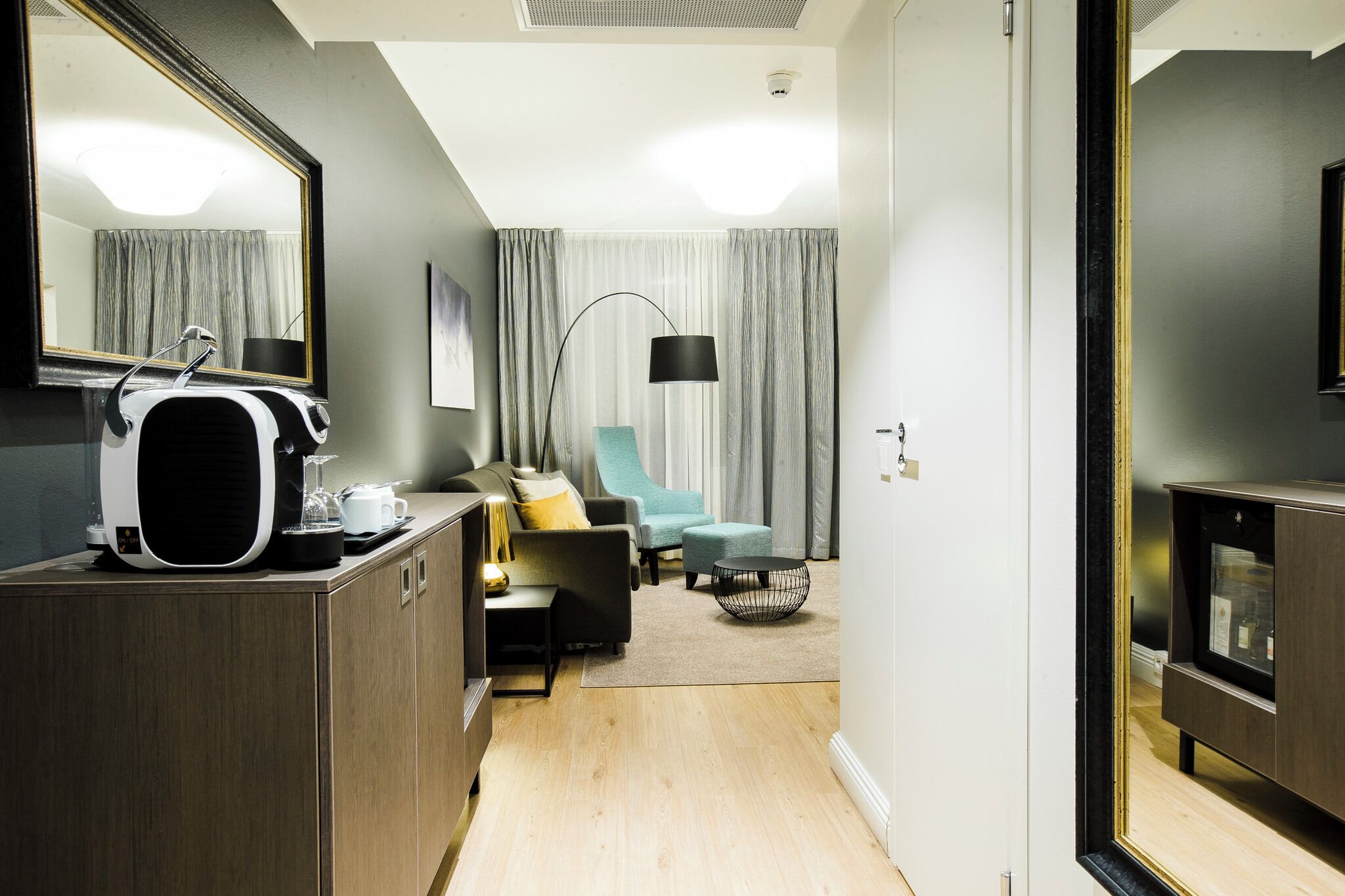A modern hotel room with a wooden cabinet, a flat-screen TV, a sofa, a chair, a floor lamp, and a large mirror.