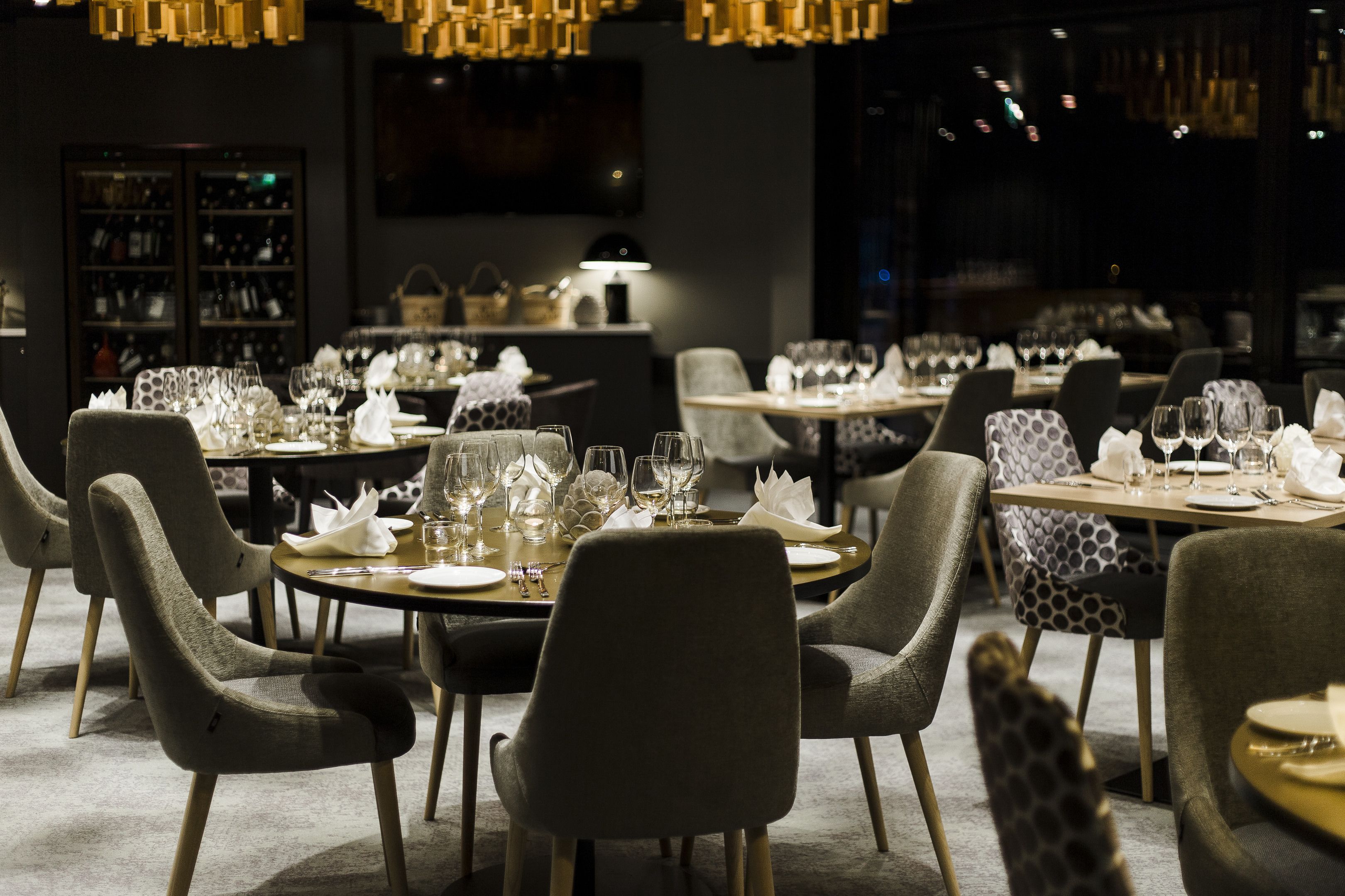 Un interior de restaurante con mesas redondas, sillas tapizadas en gris y una cálida iluminación ambiental.