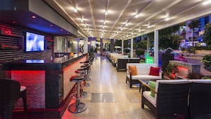 A modern bar area with a long counter, bar stools, and a television screen. There are outdoor seating areas with cushions and a view of the street. - Lonicera City Hotel (Alanya)