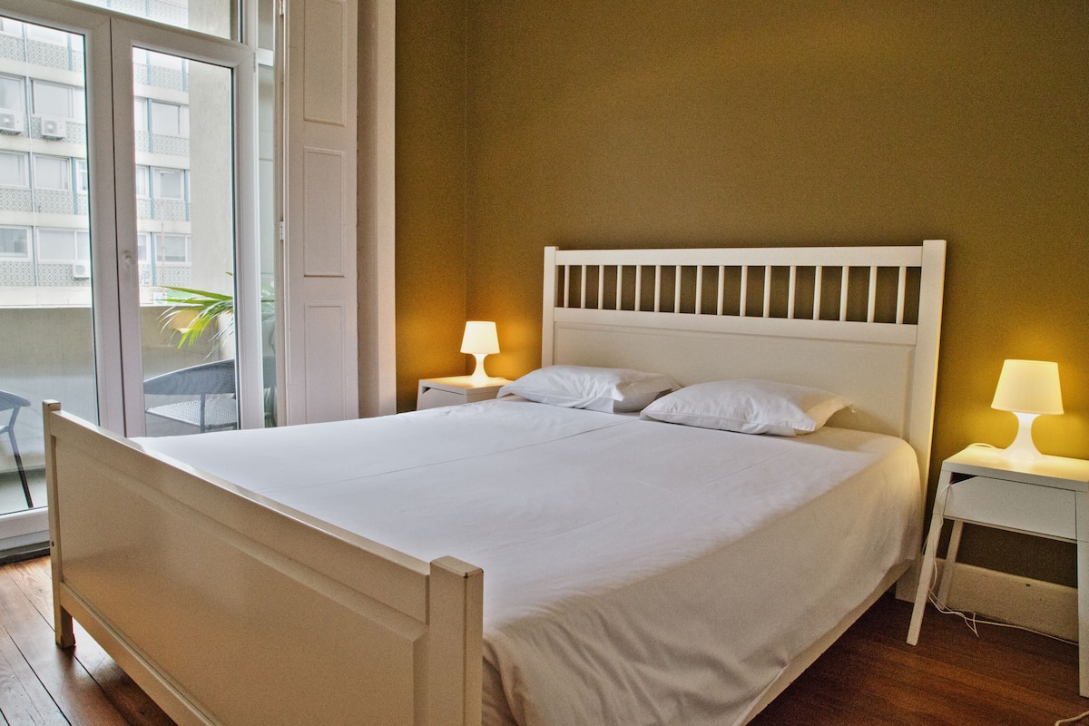 A neatly made bed with white linens in a room with wooden flooring and a warm wall color.