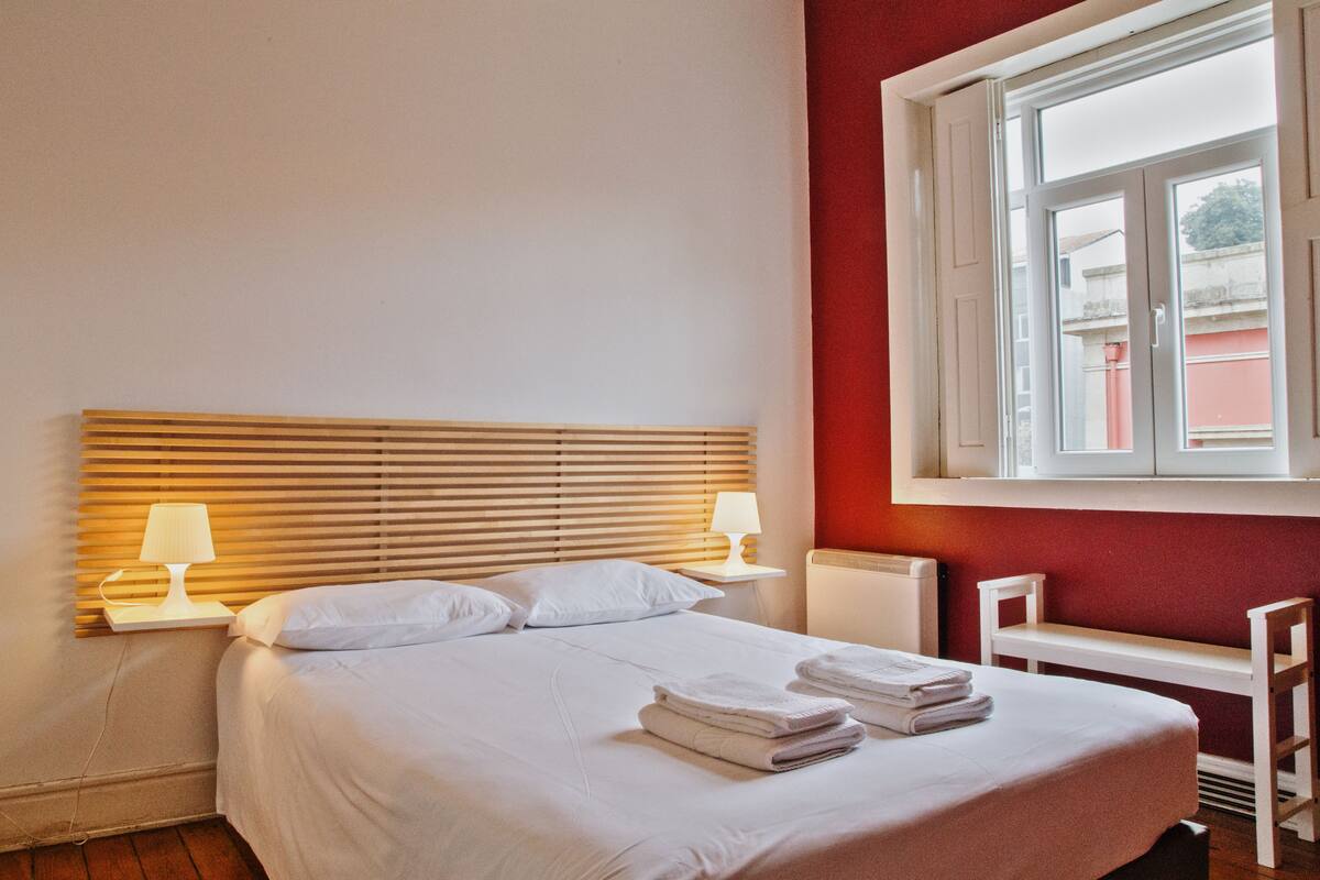 A neatly made bed with white linens and towels, a wooden headboard, and a window with blinds.
