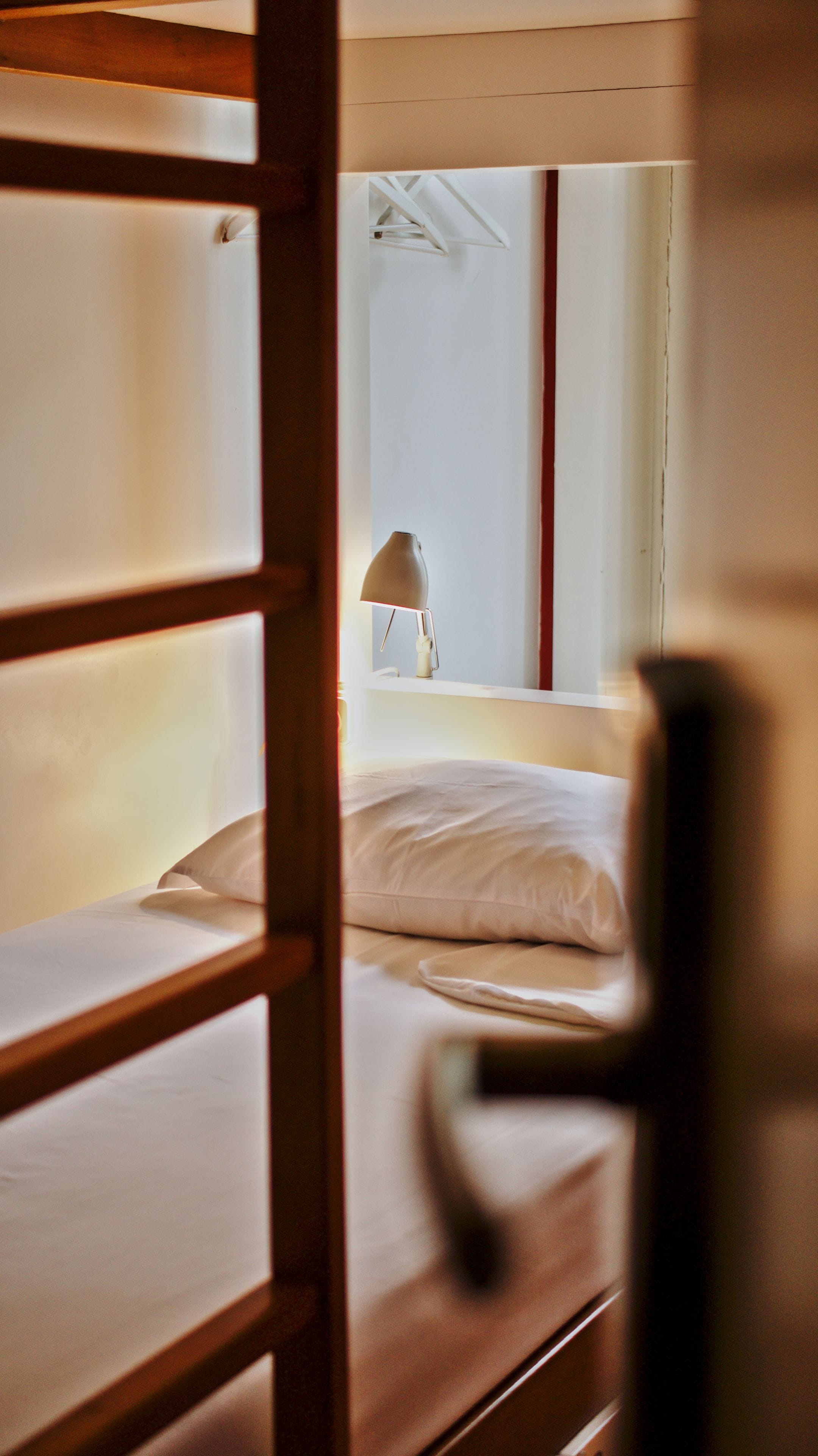 Una cama litera con mesita de noche y lámpara, una ventana con vistas al cielo, y un marco de puerta de madera.