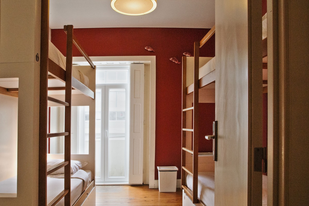 A narrow hallway with bunk beds on either side, a small white trash bin, and a door leading to a bright room.