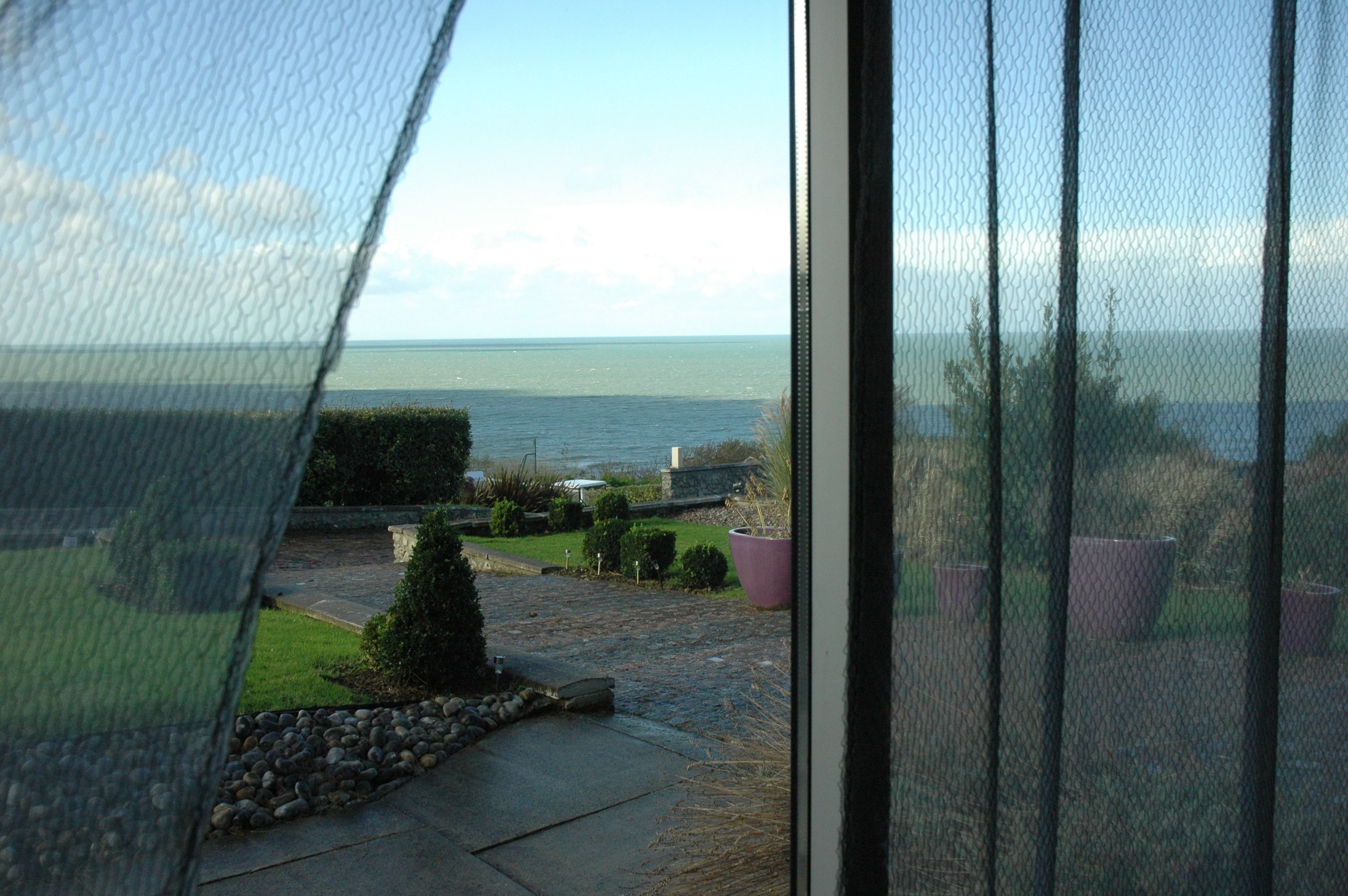 A view through a window with a mesh screen, overlooking a garden with a pathway, potted plants, and a body of water in the distance.