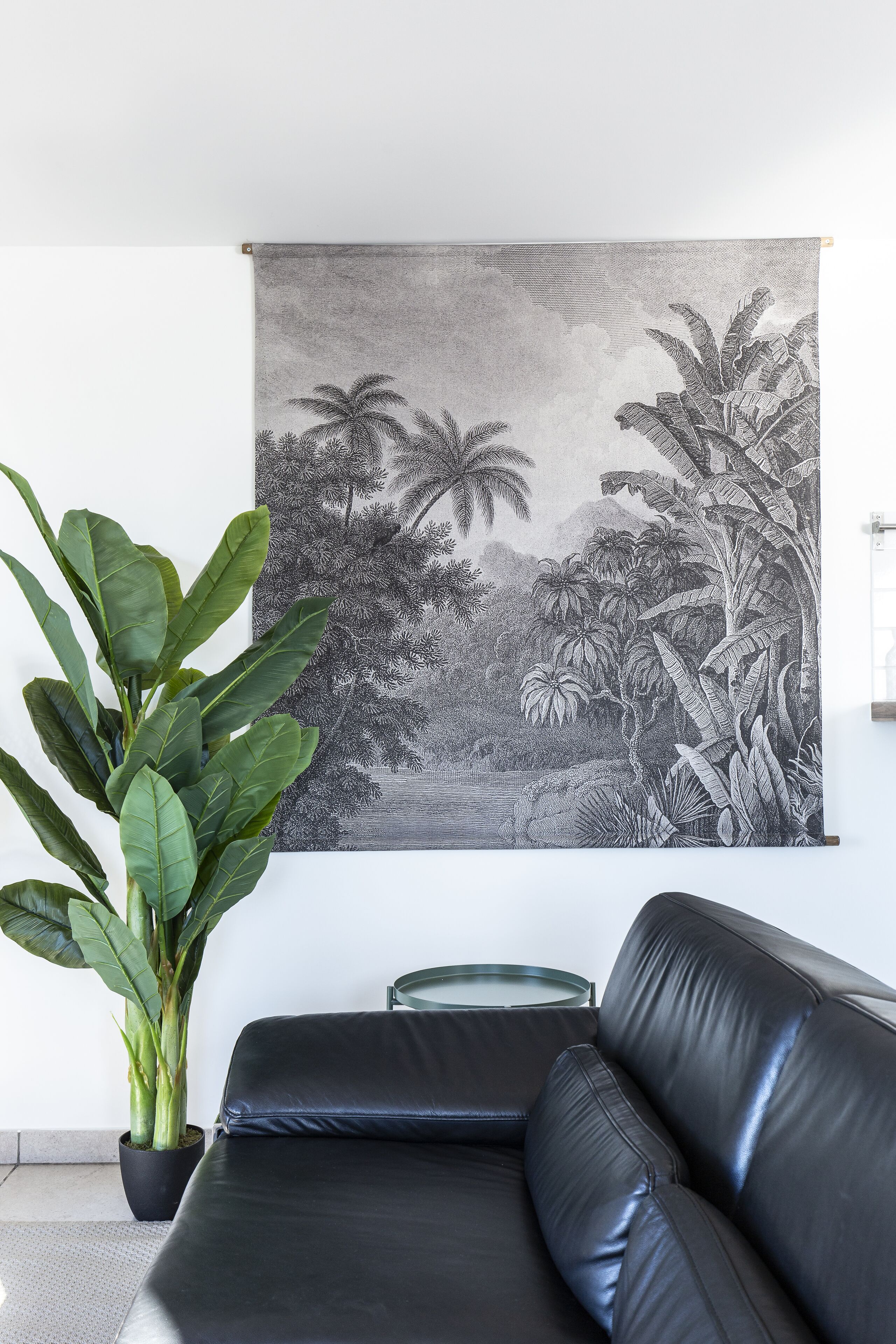 A black leather sofa, a large potted plant, and a black and white tropical landscape painting on the wall.