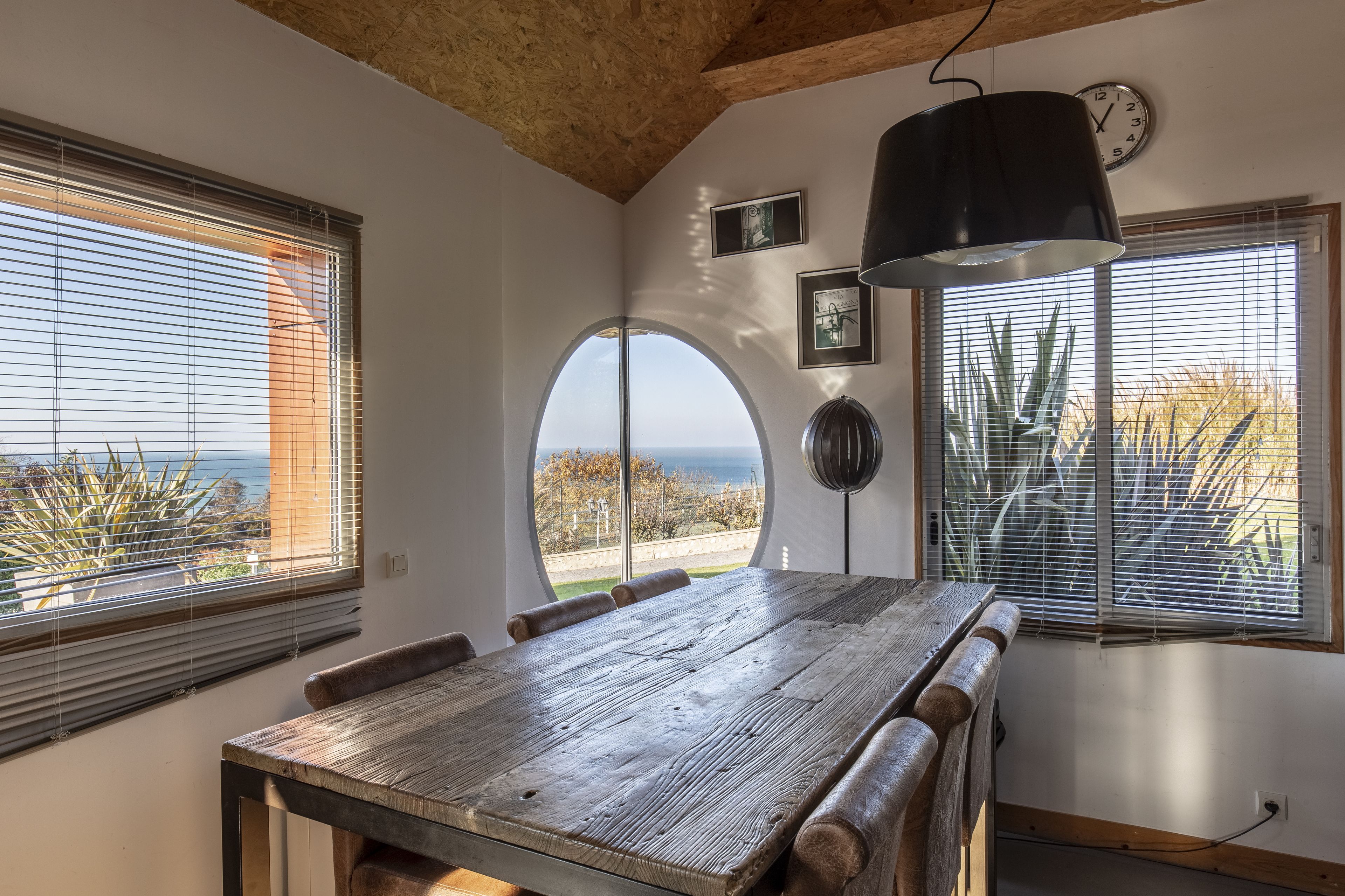 Un coin repas avec une grande table en bois, deux fenêtres équipées de stores, une horloge et des tableaux encadrés accrochés au mur.