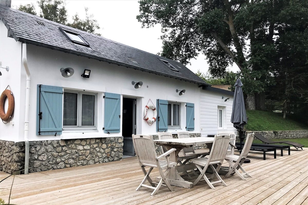 A white building with blue shutters, a stone foundation, and a wooden deck with a table and chairs.