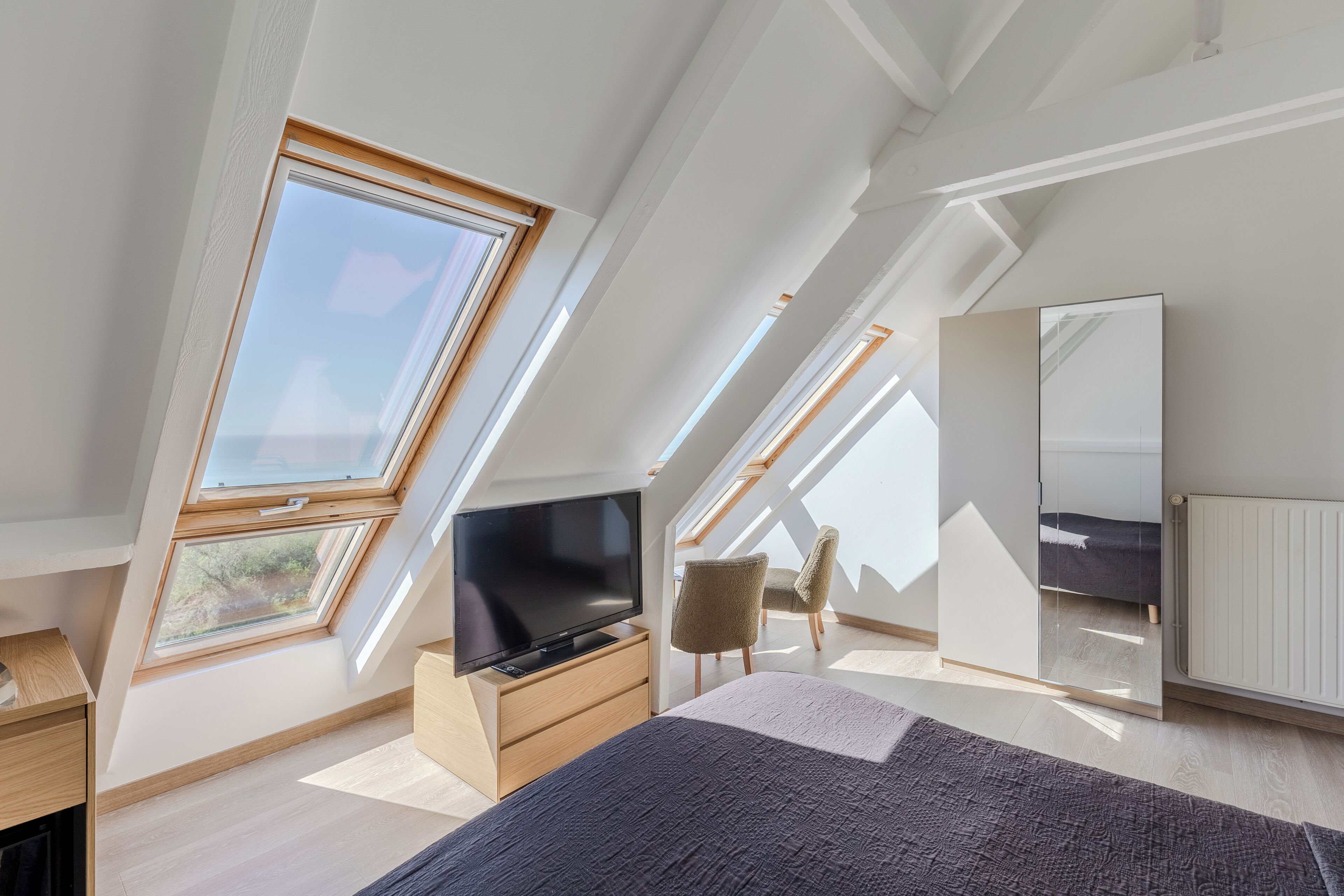 A modern loft bedroom with skylights, a bed, a TV, a dresser, and a chair.