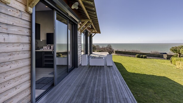 Una casa moderna con fachada de madera, grandes puertas correderas de vidrio que dan a una cocina y una terraza con vistas a un área verde y al mar.