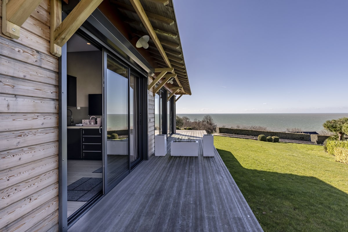 A modern house with a wooden exterior, large sliding glass doors leading to a kitchen, and a deck overlooking a grassy area and the sea.