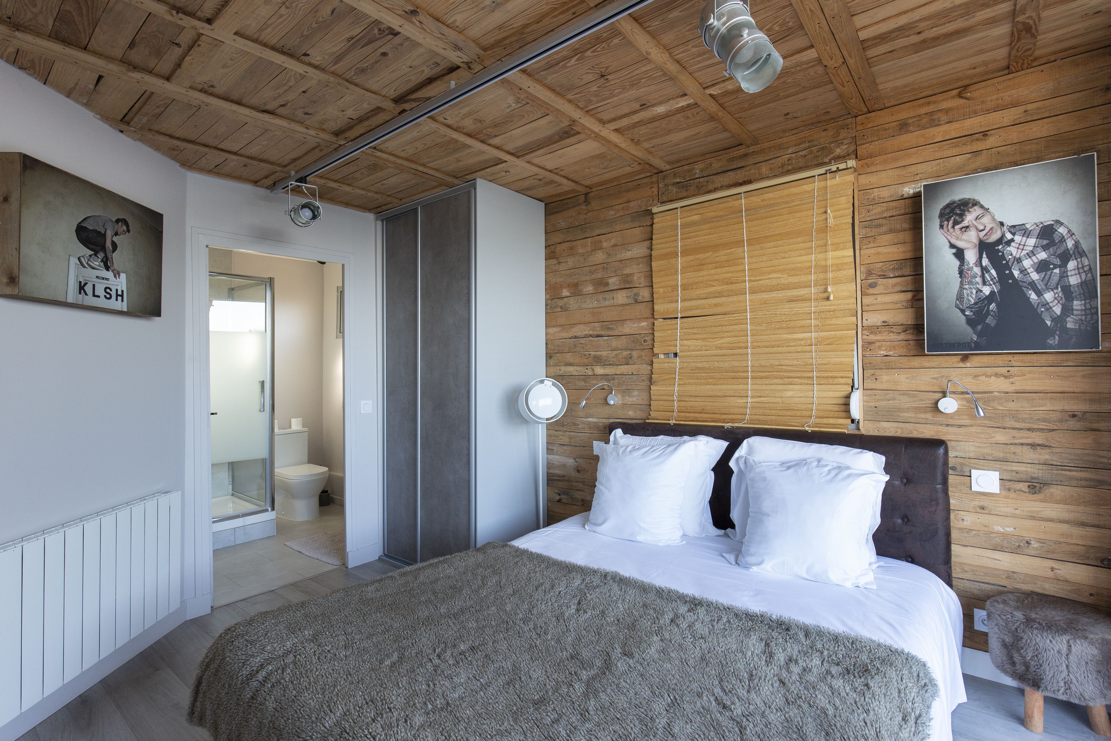 Une chambre avec un plafond en bois, un lit avec une literie blanche, une tête de lit en bois et une grande fenêtre avec des stores.