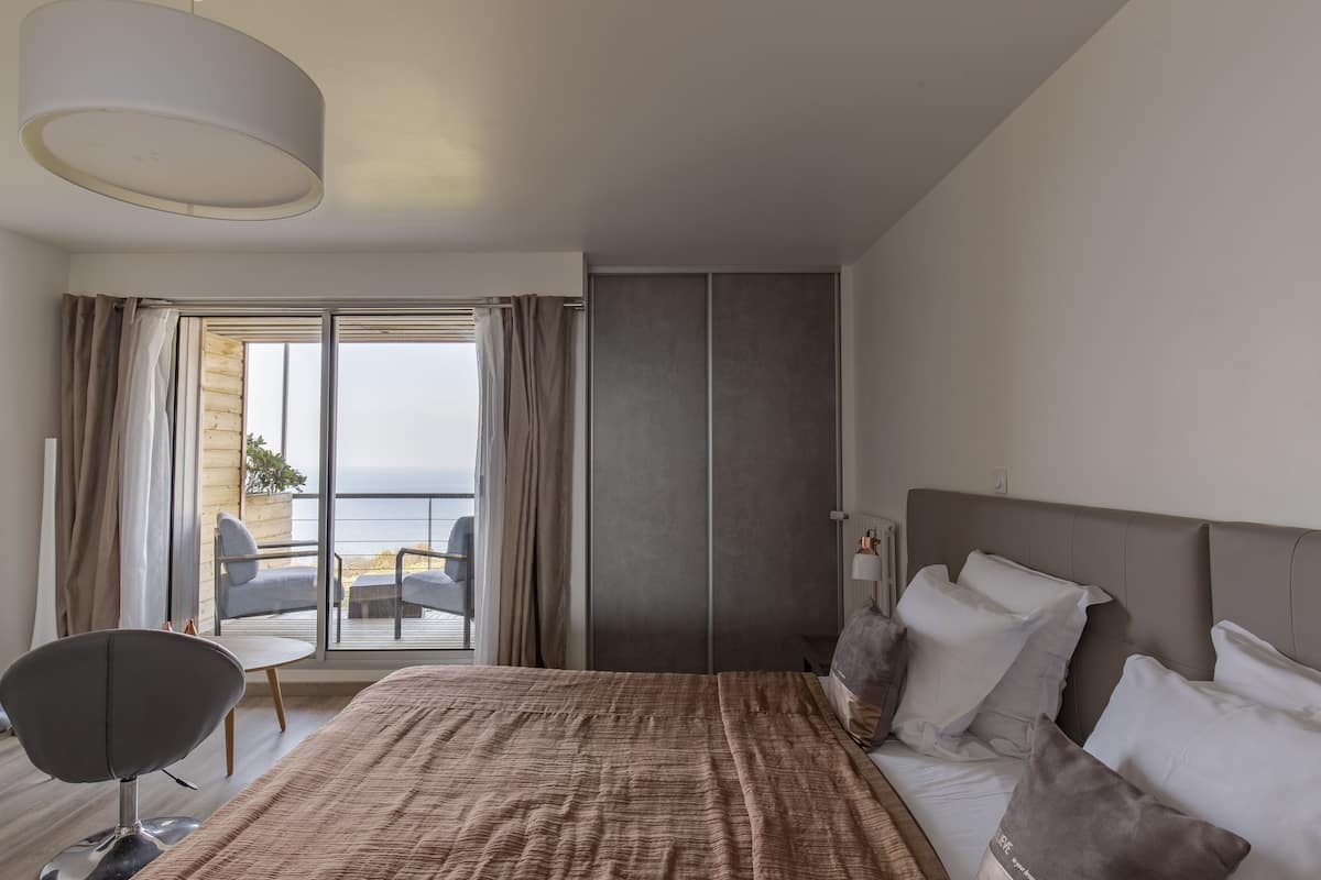 A modern hotel room with a large bed, a seating area with a chair, a view of the sea, and a sliding door closet.