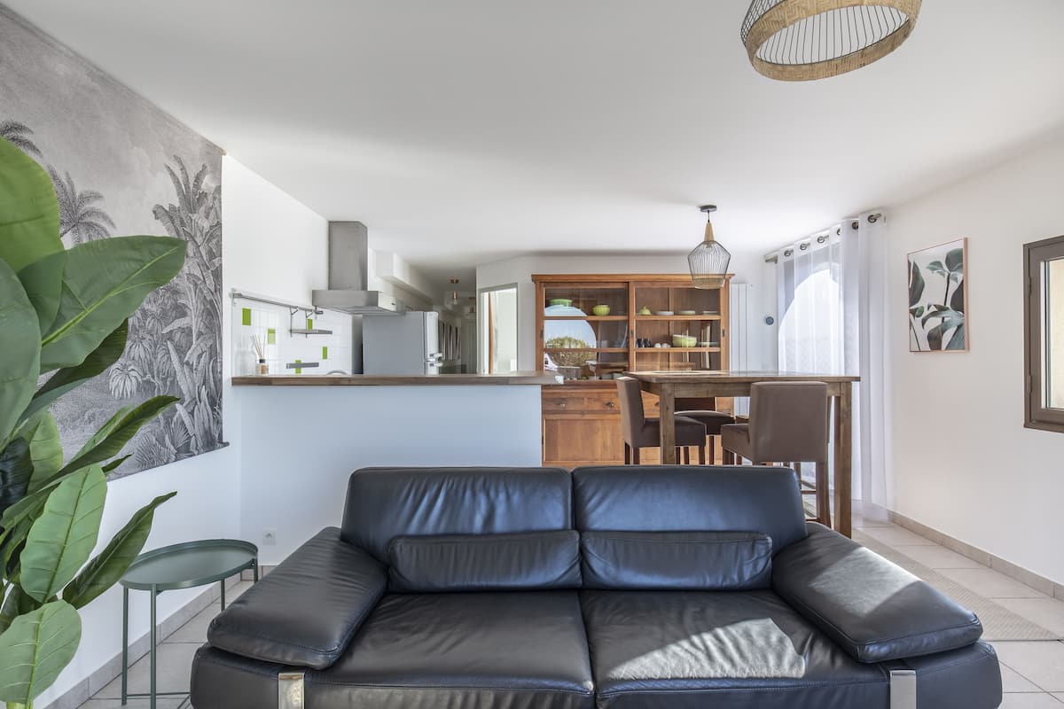 A modern living room with a black leather sofa, a dining area with a wooden table and chairs, and a kitchen with white cabinets and a hanging light fixture.