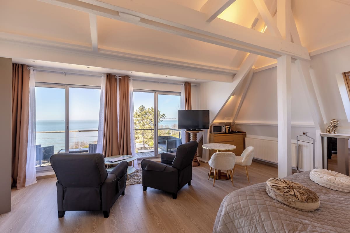 A modern hotel room with a large window, a flat-screen TV, a dining area, and a cozy bed.
