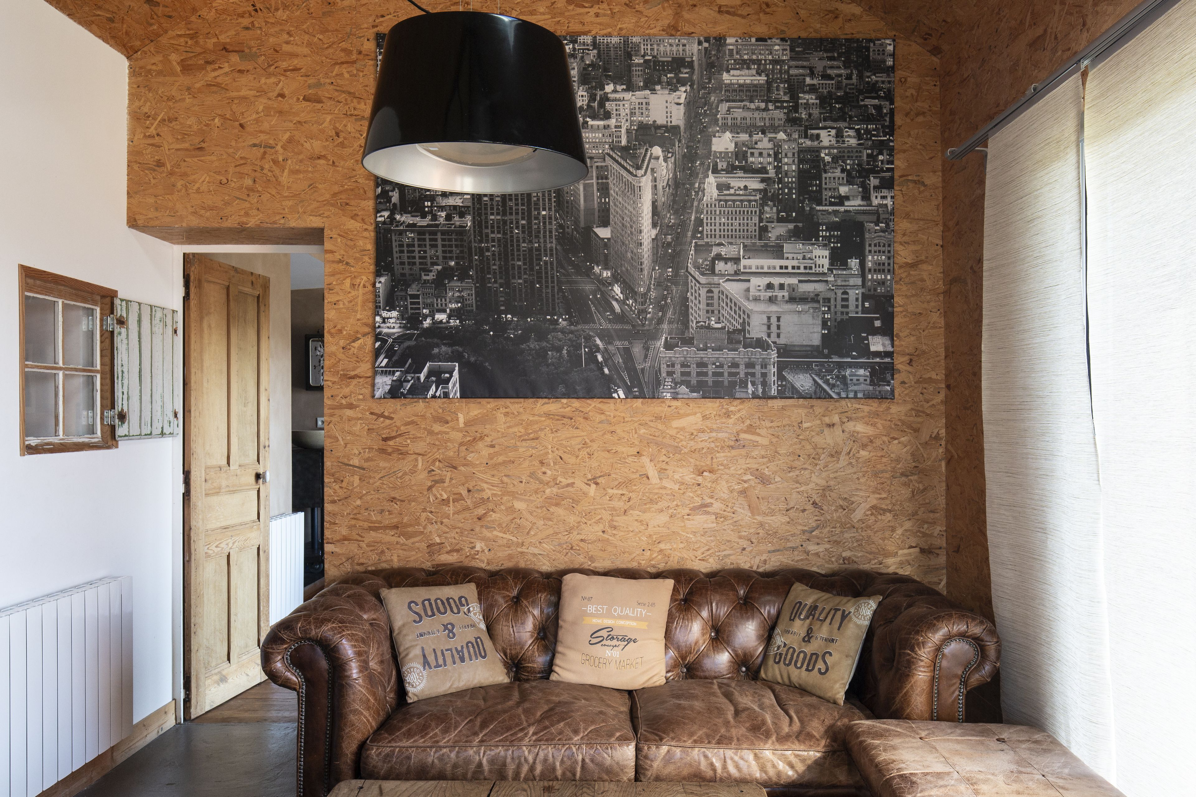 A brown leather sofa with three cushions, a black pendant lamp, and a cityscape poster on a wooden wall.