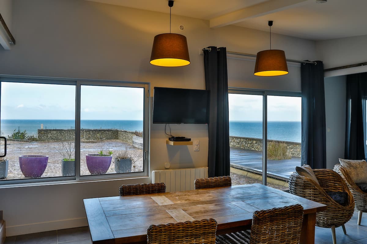 A dining area with a view of the sea, a table, chairs, and a TV mounted on the wall.