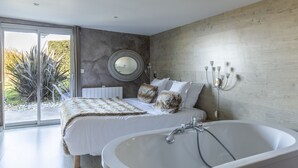 Un dormitorio moderno con una cama grande, un espejo redondo, una bañera y vistas a un jardín.