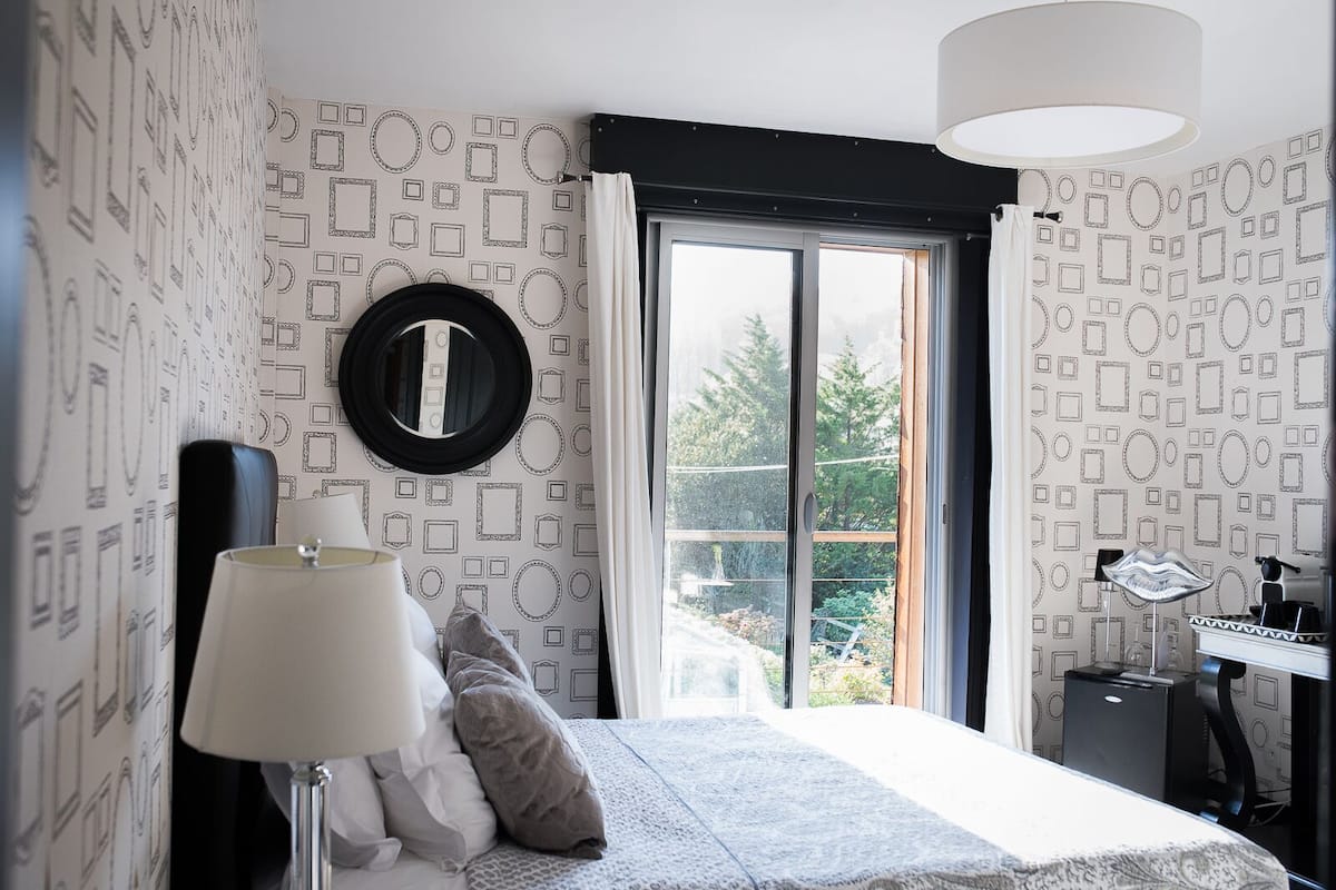 A bedroom with a bed, a nightstand, a lamp, a mirror, and a window with a view of greenery.